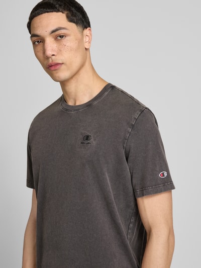 Męski model nosi T-Shirt mit Label-Stitching und Rundhalsausschnitt od CHAMPION, bawełna, krój regular fit, długość tyłu w rozmiarze M 67 cm, kolor szary, gładka tekstura, widok na górną część ciała z przodu.