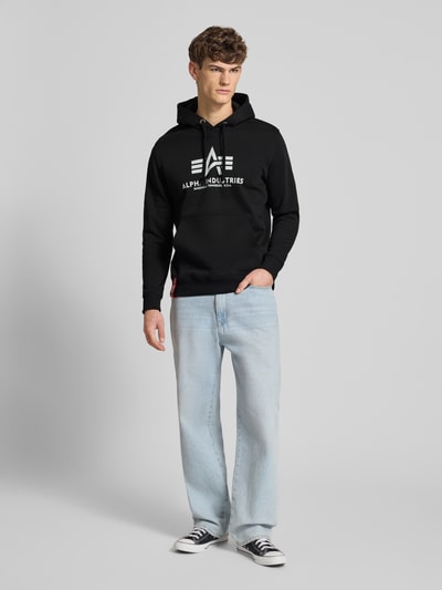 Männermodel trägt Hoodie mit Label-Print von Alpha Industries in Schwarz aus Baumwollmischung, unifarbenes Design, Ganzkörperansicht von vorne.