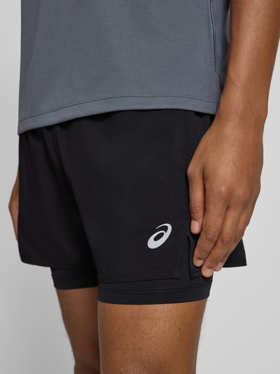 Männermodel trägt schwarze Shorts im 2-in-1-Look mit Logo von ASICS, elastischer Bund, Regular Fit, Nahaufnahme Oberschenkel.