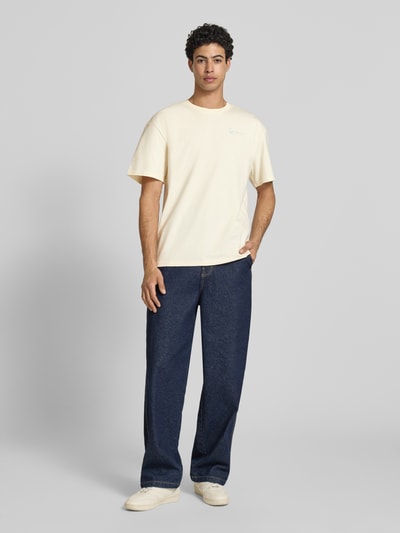 Männermodel trägt Oversized T-Shirt mit rückseitigem Print von KARL KANI aus Reine Baumwolle, Label-Stitching, Rundhalsausschnitt, Rückenlänge 73 cm, gerade geschnitten, in Creme mit glatter Textur, Ganzkörperansicht von vorne.