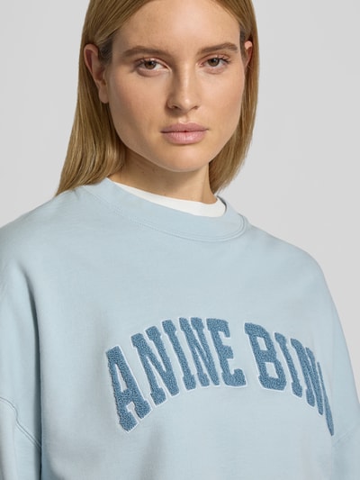Frauenmodel trägt Oversized Sweatshirt mit Logo-Detail von Anine Bing in Hellblau aus reiner Baumwolle, Oversized-Fit, Rundhalsausschnitt, elastische Abschlüsse, Nahaufnahme Oberkörper.