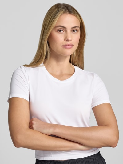 Kobiece modelka nosi biały damski t-shirt z elastycznej mieszanki materiałów, krój krótki, okrągły dekolt, Model 'Feather' od Fabletics, długość tyłu w rozmiarze S: 45 cm, ujęcie z przodu, zbliżenie na górną część ciała.