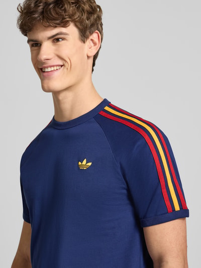 Męski model ma na sobie T-Shirt mit Label-Stitching od adidas Originals, czysta bawełna, krój regular fit, granatowy kolor z kolorowymi paskami na ramionach, wyhaftowane logo, okrągły dekolt, ściągacze, długość tyłu w rozmiarze M: 74 cm. Nahaufnahme górnej części ciała.