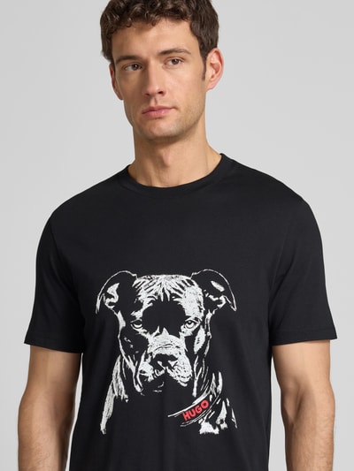 Männermodel trägt Regular Fit T-Shirt aus reiner Baumwolle Modell 'DUBROX' von HUGO, schwarz, unifarben, Motiv-Print, Nahaufnahme Oberkörper.