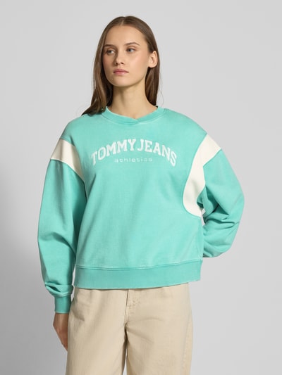 Vrouwenmodel draagt Boxy Fit Sweatshirt uit katoenmix model 'VARSITY CREW' van Tommy Jeans in two tone optic, met label stitching en geribde contraststrepen; ronde hals, lange mouwen, sweatmateriaal. Vooraanzicht borst close-up.