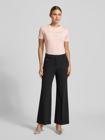 Frauenmodel trägt T-Shirt mit Strasssteinbesatz Modell 'SIENA' von Guess, hellrosa mit Logo-Print, Baumwoll-Mix, Geradeform, Ganzkörperansicht von vorne.