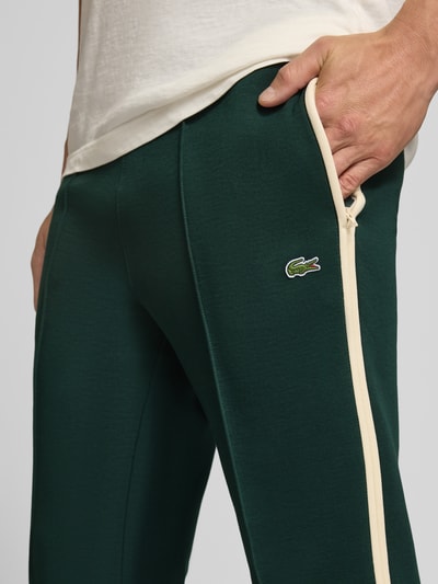 Männermodel trägt Regular Fit Jogpants aus Baumwoll-Mix von Lacoste in Dunkelgrün mit beigem Streifen, Nahaufnahme Oberschenkelansicht. Herren Weite Hose von Lacoste
