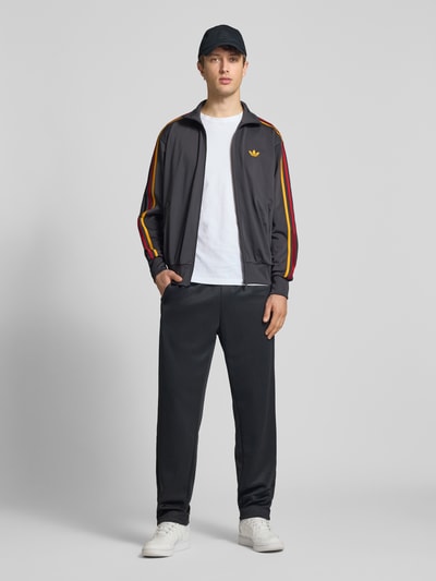 Männermodel trägt Regular Fit Trainingsjacke mit mehrfarbigen Kontraststreifen Modell 'FIREBIRD TT' von adidas Originals, Optik: Unifarben, Passform: Regular-Fit, Logo-Stitching, Stehkragen, Langarm, Ärmelabschluss: gerippt, Rumpfabschluss: gerippt, seitliche Reißverschlusstasche, dunkle Grundfarbe mit auffälligen Streifen, glatte Textur, Ganzkörperansicht von vorne.