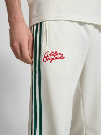 Männermodel trägt Sweatpants mit Reißverschlusstaschen und Logo-Stitching von adidas Originals in Weiß mit grünen Streifen, Material-Mix mit Baumwoll-Anteil, Lockerer-Schnitt, Elastischer-Bund, Detailaufnahme Oberschenkel.