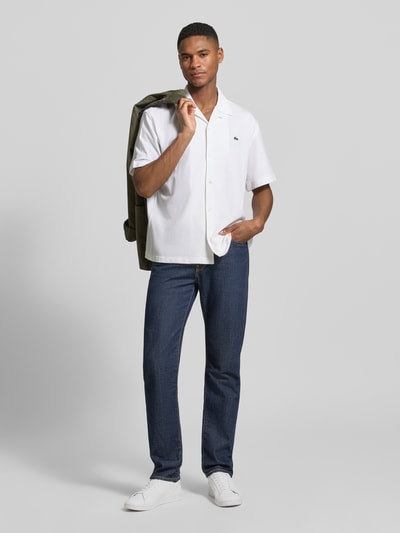 Männermodel trägt Relaxed Fit Freizeithemd aus reiner Baumwolle von Lacoste, unifarben weiß mit Kentkragen und kurzem Arm, Ganzkörperansicht von vorne.