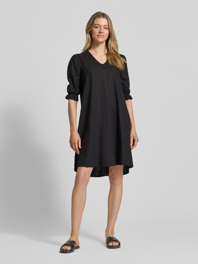 Frauenmodel trägt knielanges Kleid aus Leinen-Viskose-Mix Modell 'FALAKKA' von B.Young in Schwarz, gerader Schnitt, V-Ausschnitt, Rüschen, Ganzkörperansicht von vorne. Rückenlänge 95 cm, Brustweite 38 cm, Ärmellänge 39 cm.