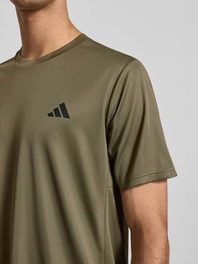 Männermodel trägt T-Shirt mit Label-Print von adidas Training in Oliv, Lockerer-Schnitt, gerippter Rundhalsausschnitt, Nahaufnahme Oberkörper. Rückenlänge bei Größe M: 70 cm.