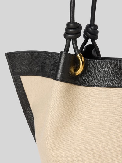 Produktfoto, Shopper mit Label-Detail Modell 'FINN CANVAS' von Coccinelle aus beigefarbenem Canvas mit schwarzen Lederelementen und goldfarbenem Metall, sichtbare grobe Textur, Detailaufnahme, Hauptfach mit Druckknopfverschluss, lange Tragegriffe, Reißverschlussfach innen