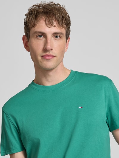 Männermodel trägt Regular Fit T-Shirt aus reiner Baumwolle von Tommy Jeans, grün, Logo-Stitching, Nahaufnahme Oberkörper.