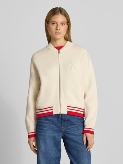 Frauenmodel trägt Oversized Bomberjacke aus Baumwoll-Mix mit Woll-Anteil Modell 'ICONIC A' von Armedangels, unifarben creme mit roten Rippstrick-Details, Zweiwege-Reißverschluss und Logo-Stitching, Ganzkörperansicht von vorne.