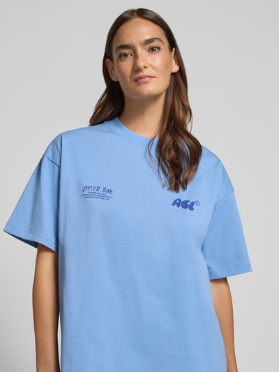 Modelka kobieca nosi damski t-shirt z bawełny o kroju relaxed fit model 'Oyster Bar' od ANOTHER COTTON LAB z nadrukiem z logo, niebieski, okrągły dekolt, długość tyłu w rozmiarze S: 61 cm, z przodu, zbliżenie na górną część ciała.
