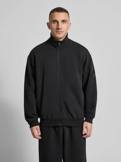 Männermodel trägt Loose Fit Sweatjacke mit Modal und Baumwoll-Anteil von adidas Sportswear, Optik: Unifarben, schwarze Farbe, Struktur: glatter Sweat, Logo-Detail am Ärmel, Stehkragen, Ärmelabschluss und Rumpfabschluss: Gerippt, Verschluss: Reißverschluss, seitliche Eingrifftaschen, Materialverarbeitung: Material-Mix, Ganzkörperansicht von vorne.