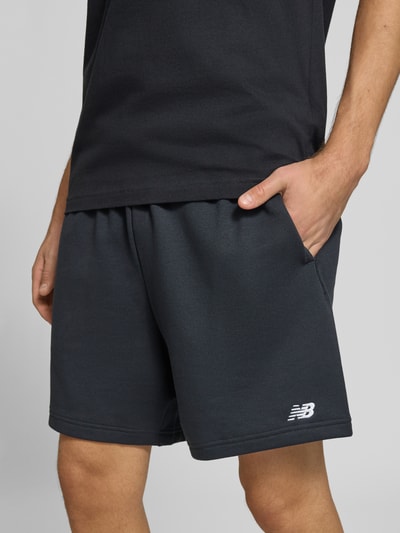 Männermodel trägt schwarze Sweatshorts aus Baumwoll-Mix Modell 'FRENCH' von New Balance, elastischer Bund, Logo-Prägung, Nahaufnahme Oberkörper, Kontext: Herren Sweatshorts, gerade Passform, Gesäßtasche, Bundweite 40 cm, Innenbeinlänge 17 cm.