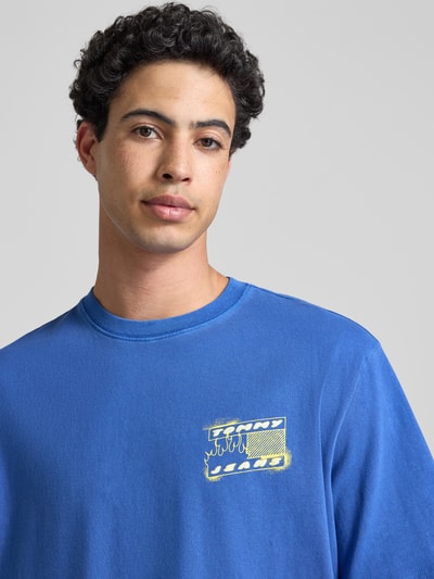 Männermodel trägt Regular Fit T-Shirt aus reiner Baumwolle Modell '90s' von Tommy Jeans in Blau mit Label Print, Nahaufnahme Oberkörper.