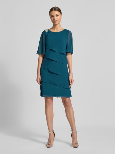 Frauenmodel trägt Relaxed Fit Kleid mit Ziersteinbesatz von Christian Berg Cocktail in Türkis, semitransparent, mit Rüschen, Ganzkörperansicht von vorne.