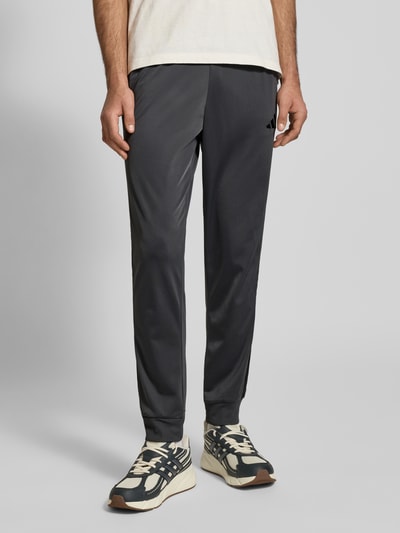 Männermodel trägt Slim Fit Trainingsanzug mit Logo-Stitching von adidas Sportswear, dunkle Farbe, elastische Abschlüsse, Nahaufnahme Unterkörper. Kontext: Herren Trainingsanzug, Stehkragen, Eingrifftaschen, Reißverschluss, Bundweite 39 cm, Rückenlänge 71 cm, Schulterbreite 47 cm, Ärmellänge 66 cm, Innenbeinlänge 76 cm.