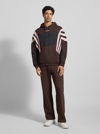 Männermodel trägt Hoodie mit Kapuze von adidas Originals in Braun mit rosa Streifen, Känguru-Tasche, Gerippte-Abschlüsse, Lockerer-Schnitt, Ganzkörperansicht von vorne. Rückenlänge bei Größe M: 71 cm, Ärmellänge bei Größe M: 66 cm, Label-Stitching.