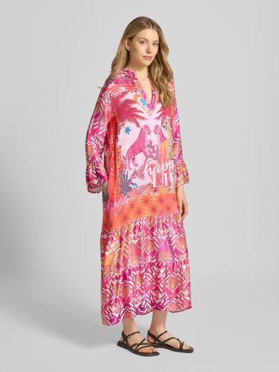 Frauenmodel trägt Tunikakleid aus Viskose mit Trompetenärmeln von miss goodlife, leuchtende Pink-Orange-Töne, Lockerer-Schnitt, Modell 'Tiger Heart', Serafino-Ausschnitt, Midikleid, auffälliges Tiger-Motiv, Volantsaum, Ganzkörperansicht von vorne, Rückenlänge bei Größe S: 130 cm, Brustweite: 52 cm, Ärmellänge: 56 cm.