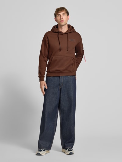 Männermodel trägt Hoodie mit Känguru-Tasche von Alpha Industries in Braun, lockerer Schnitt, Ganzkörperansicht von vorne, Baumwoll-Mix