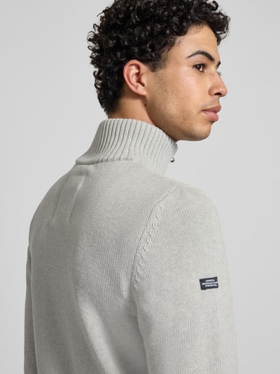 Männermodel trägt Slim Fit Pullover in imitacja dzianiny von Superdry, krój slim fit, stójka, zamek błyskawiczny, wyhaftowane logo, długość tyłu w rozmiarze M 70 cm, długość rękawów w rozmiarze M 65 cm, jasnoszary kolor, widoczne tekstury dzianiny, detailaufnahme