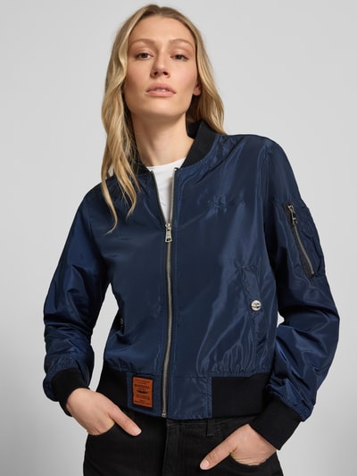 Frauenmodel trägt Bomberjacke mit Label-Patch und Reißverschlusstasche am Ärmel von Original Bombers in Dunkelblau, leicht glänzende, glatte Textur, mit Stehkragen, Reißverschluss, Logo-Stitching, Seitentaschen mit Druckknopfverschluss und elastischen Abschlüssen, Lockerer-Schnitt, Nahaufnahme Oberkörper. Rückenlänge 53 cm, Schulterbreite 37 cm, Brustweite 48 cm, Ärmellänge 63 cm bei Größe S.