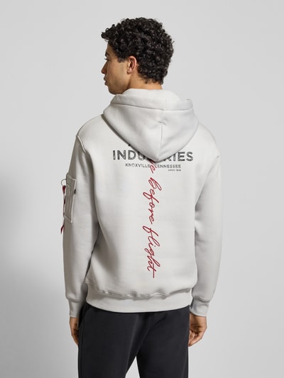 Männermodel trägt Hoodie mit Label-Details von Alpha Industries in Off-White, Baumwoll-Mix, Reißverschlusstasche am Ärmel, Lockerer-Schnitt, Kapuze, Känguru-Tasche, gerippte Abschlüsse, Rückenlänge bei Größe M 68 cm, Ärmellänge 67 cm, Ganzkörperansicht Rückansicht