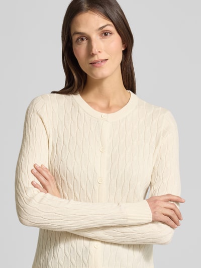 Modelka kobieca nosi Regular Fit Strickjacke mit Viskose-Anteil Modell 'FANTASTIC' od Vero Moda, kremowy kolor, faktura warkoczowy splot, długi rękaw, okrągły dekolt, przód na guziki, detal label stitching, widok na górną część ciała z przodu.