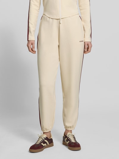 Kobiece modelka nosi Jogpants mit Logo Modell 'JASMINE' od Guess Activewear, kolor kremowy, materiał z Lyocellem, krój regular fit, logo, kieszenie, zdjęcie całego ciała z przodu.