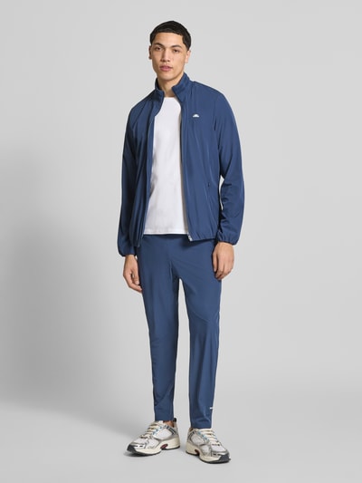 Männermodel trägt spodnie typu track pants z elastycznym pasem model 'EVARO' od Ellesse, mieszanka materiałów z dodatkiem streczu, krój regular fit, długość wewnętrzna nogawki w rozmiarze M: 74 cm, całkowity widok z przodu.