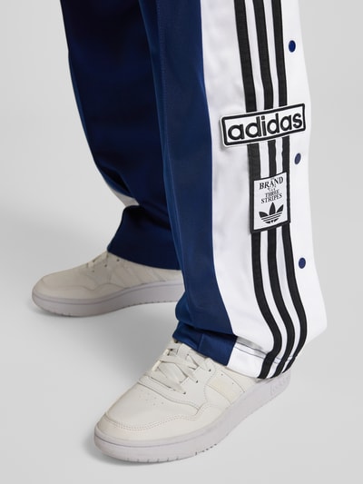 Frauenmodel trägt Straight Fit Jogginghose mit elastischem Bund Modell 'Adibreak' von adidas Originals, dunkelblau-weiß mit Ziernähten, Eingrifftaschen und Streifen; Nahaufnahme Bein.