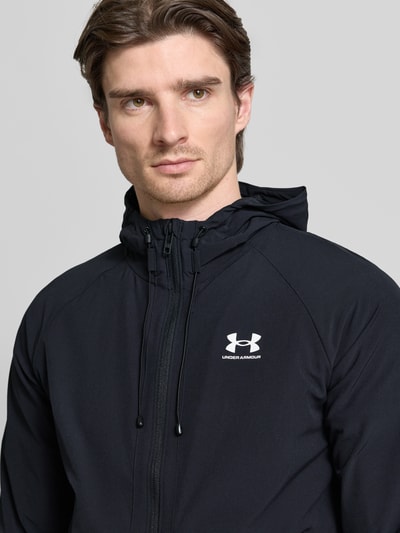 Männermodel trägt Kurtka mit Kapuze von Under Armour, mieszanka materiałów z dodatkiem streczu, luźny krój, tunel, zamek błyskawiczny, nadruk z logo, wpuszczane kieszenie, długość tyłu w rozmiarze M 68 cm, długość rękawów w rozmiarze M 66 cm, kolor czarny, prostokątne formy, gładka tekstura, nahaufnahme oberkörper.