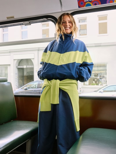 Vrouwenmodel draagt Nylon-Jacke im 90s-Stil - curated by Tinvcb van Tinvcb in blauw met limegroene strepen, regular fit, met opstaande kraag, ritssluiting, elastische boorden, ritszijzakken, ruglengte 60 cm, schouderbreedte 54 cm, borstwijdte 58 cm, mouwlengte 34 cm, totaalbeeld van voren