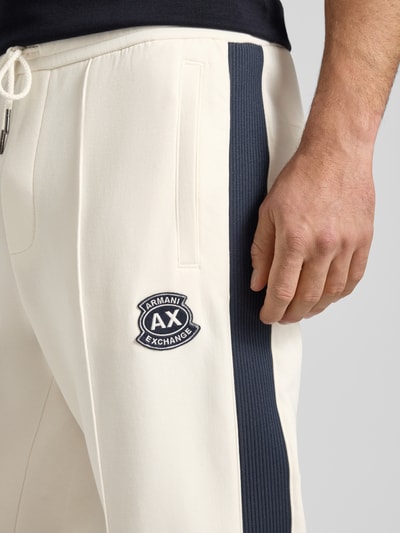 Männermodel trägt Sweatpants mit Label-Patch und elastischem Bund von ARMANI EXCHANGE, cremefarben mit schwarzem Seitenstreifen, Nahaufnahme Oberkörper. Baumwoll-Mix mit Stretch-Anteil, Relaxed-Fit, Längsnähte, Innenbeinlänge 42 cm bei Größe M.