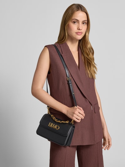 Männermodel trägt Handtasche mit Label-Applikation von Liu Jo White, schwarz mit goldener Kette und Schulterriemen, Szerokość w rozmiarze 1: 23 cm, długość: 14 cm, głębokość: 8 cm, długość paska: 110 cm, Ganzkörperansicht von vorne
