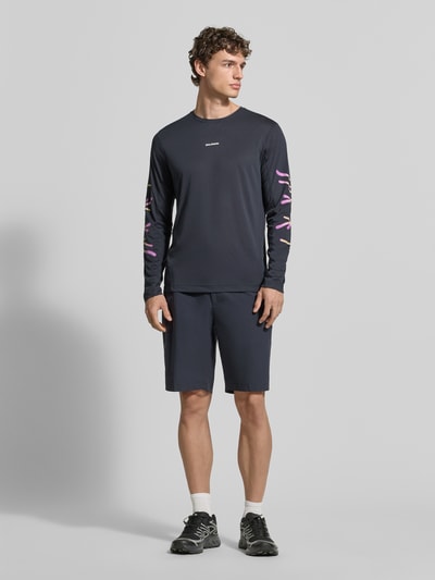 Männermodel trägt Funktionsshirt mit Logo Modell 'Kout' von Salomon, Regular Fit, Longsleeve, dunkelblau, schlichte Textur, Ganzkörperansicht von vorne