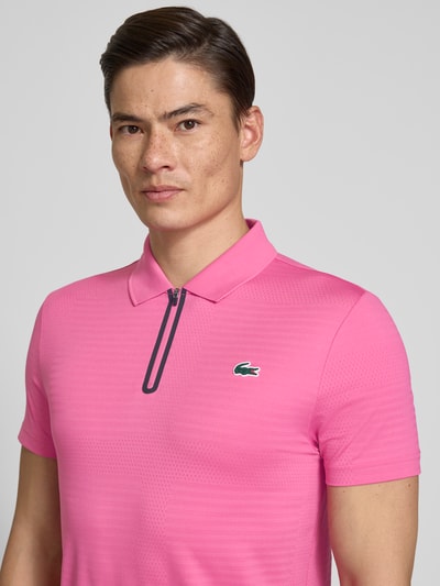 Męski model nosi Slim Fit Poloshirt aus schnelltrocknendem Material von Lacoste Sport, różowy kolor, logo, krótki rękaw, krój slim fit, szybkoschnący, właściwości zapobiegające powstawaniu zapachów, jednolita faktura; zbliżenie na górną część ciała z przodu.