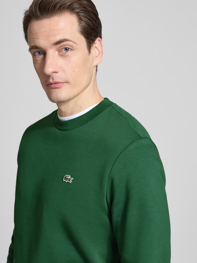 Mannenmodel draagt Regular Fit Sweatshirt uit katoenmix van Lacoste in effen groene look met labelpatch, ronde hals, lange mouw, sweat materiaal, detailopname bovenlichaam.