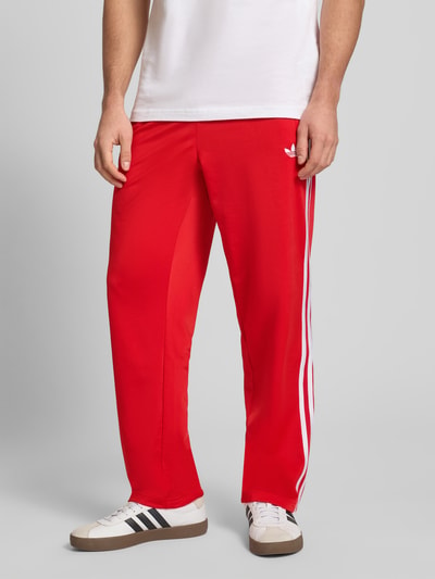Mężczyzna nosi Sweatpants mit elastischem Bund od adidas Originals, luźny krój, czerwona tkanina, elastyczny pas, naszywka z logo, kieszenie na zamek, długość wewnętrzna nogawki w rozmiarze M: 79 cm, widok całej sylwetki z przodu.