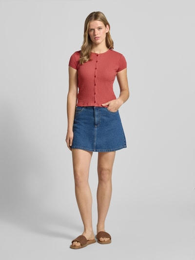 Frauenmodel trägt Slim Fit T-Shirt mit durchgängiger Knopfleiste Modell 'AJOUR' von REVIEW aus Reine Baumwolle, Rundhalsausschnitt, kurzärmelig, in Altrosa mit feiner Rippenstruktur, kombiniert mit Jeansrock, Ganzkörperansicht von vorne.