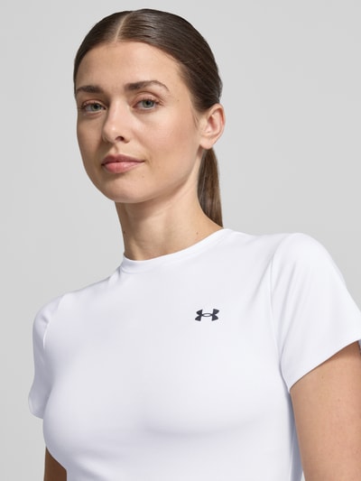 Kobiece modelka nosi T-Shirt mit Rundhalsausschnitt und Logo od Under Armour, biały, krój slim fit, materiał z domieszką stretchu, logo, długość pleców w rozmiarze S 49 cm, zbliżenie na górną część ciała z przodu.