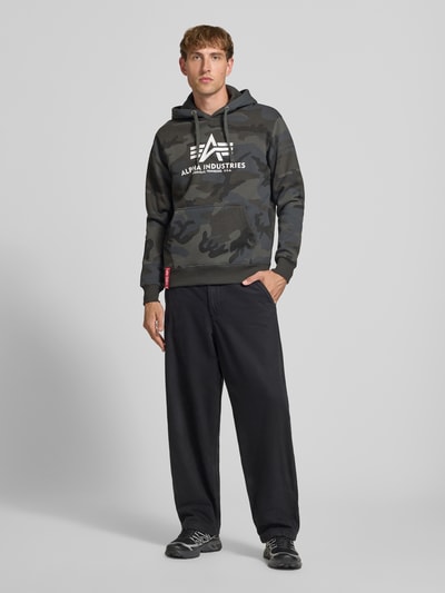 Männermodel trägt Regular Fit Hoodie aus Baumwoll-Mix mit Logo-Print von Alpha Industries in Camouflage, Kapuze, Kängurutasche, Ganzkörperansicht von vorne.
