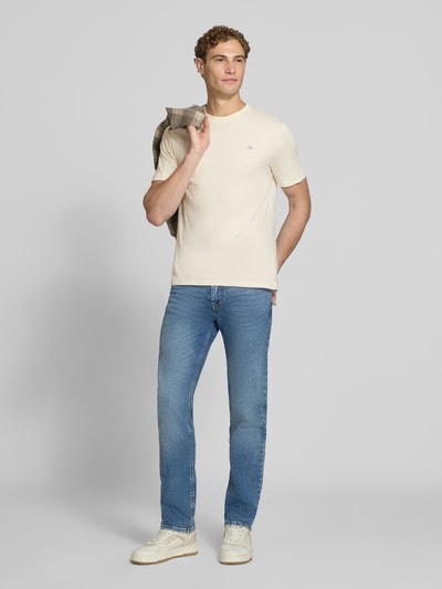Männermodel trägt Regular Fit T-Shirt aus reiner Baumwolle von Scotch & Soda in Beige, dezentes Logo-Stitching, Jeans, Ganzkörperansicht von vorne. Rückenlänge bei Größe M: 61 cm.