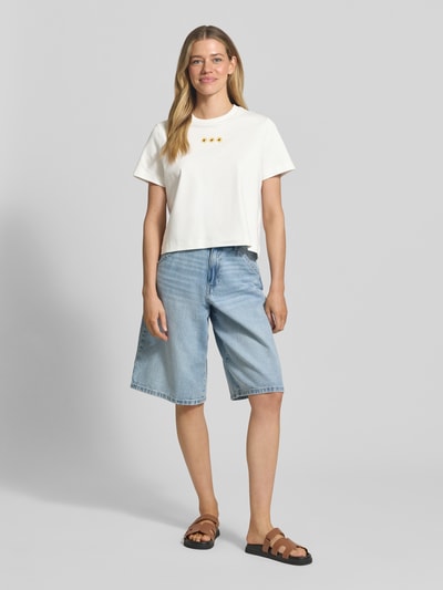 Frauenmodel trägt Regular Fit T-Shirt aus reiner Baumwolle Modell 'SARA' von Vero Moda, unifarben, mit Logo-Stitching, Rundhals, kurzarm, Jersey-Textur, kombiniert mit hellblauer Jeansbermuda, neutrale Farbtöne, Ganzkörperansicht von vorne.