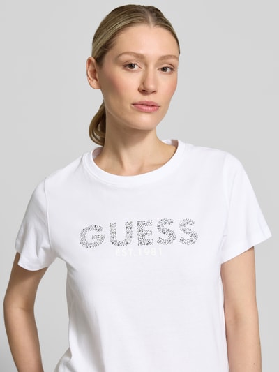 Frauenmodel trägt T-Shirt mit Label-Print und Strasssteinbesatz von Guess aus reiner Baumwolle, Straight Fit, Rundhalsausschnitt, Nahaufnahme Oberkörper.
