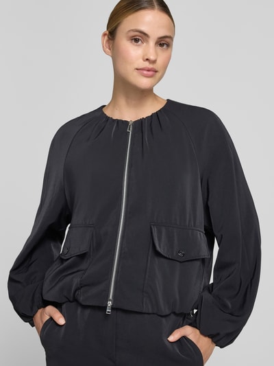 Frauenmodel trägt Blouson mit Zweiwege-Reißverschluss und Pattentaschen von Marc Cain in Regular-Fit, schwarze Farbe, Nahaufnahme Oberkörper, Kontext: Damen Blouson, Rundhalsausschnitt, Rückenlänge 56 cm, Brustweite 60 cm, Ärmellänge 62 cm bei Größe 36.